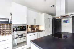 Kuchynský nábytok s povrchom v elegantnej bielej farbe s kontrastne tmavou pracovnou doskou doplňuje ostrovček s varnou doskou a digestorom v tom istom vyhotovení