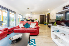 Pohľad do hlavnej obytnej miestnosti domu odhaľuje obytnú sekciu v popredí so sedacou súpravou, nasleduje jedálenský stôl pre celú rodinu a biela kuchyňa. V celom dome sú zvýšené dvere, aby vzduch ľahšie cirkuloval vo vnútornom priestore